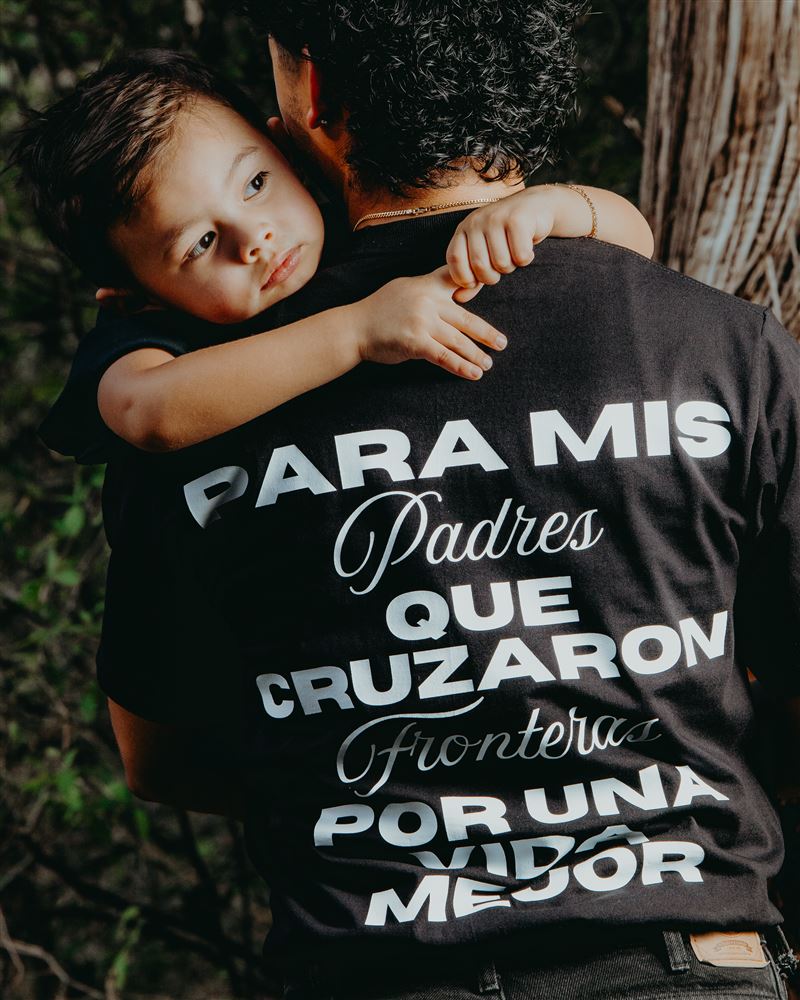 Man in a black Destino tee carrying a young boy on his shoulder; the back of the shirt reads Para mis padres que cruzaron fronteras por una vida mejor