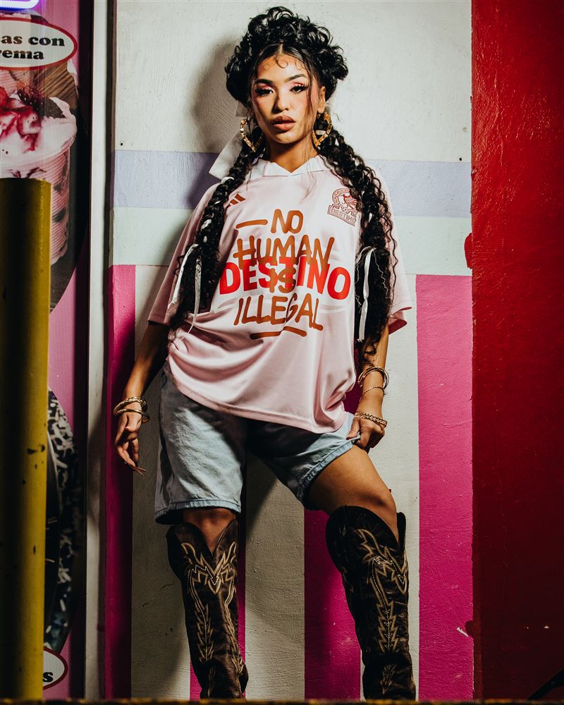 Woman in the pink Destino jersey and western boots against a pink and white striped wall at night