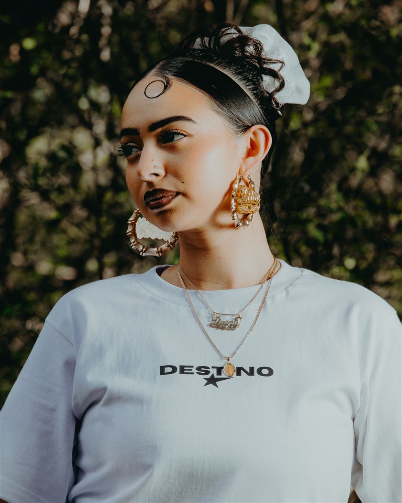 Woman in a white Destino tee with gold hoops and a name necklace, lit by a soft key