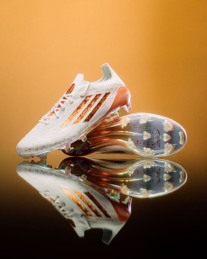adidas F50 Elite boots in white and gold on a reflective surface, warm backlight